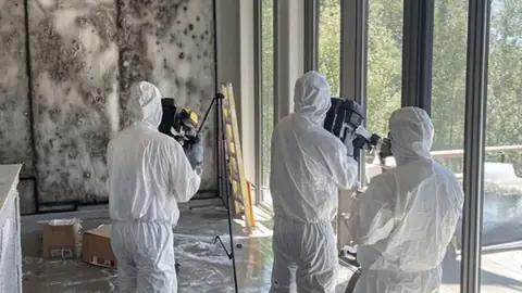 Crew in protective suits with equipment in a room with mold-stained walls